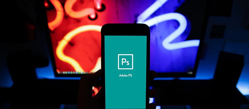 Hand holding a phone with Adobe Photoshop app open, colorful neon background displayed on monitor.