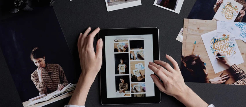 Hands working on a tablet with printed photos and design sketches laid out on a dark table